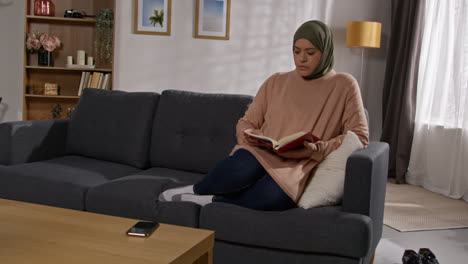 Muslimische-Frau-Mit-Hijab-Sitzt-Zu-Hause-Auf-Dem-Sofa-Und-Liest-Oder-Studiert-Den-Koran